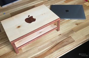 A pine laptop riser with inlaid Sapele Apple logo sitting next to an Apple laptop. Links to the corresponding product page