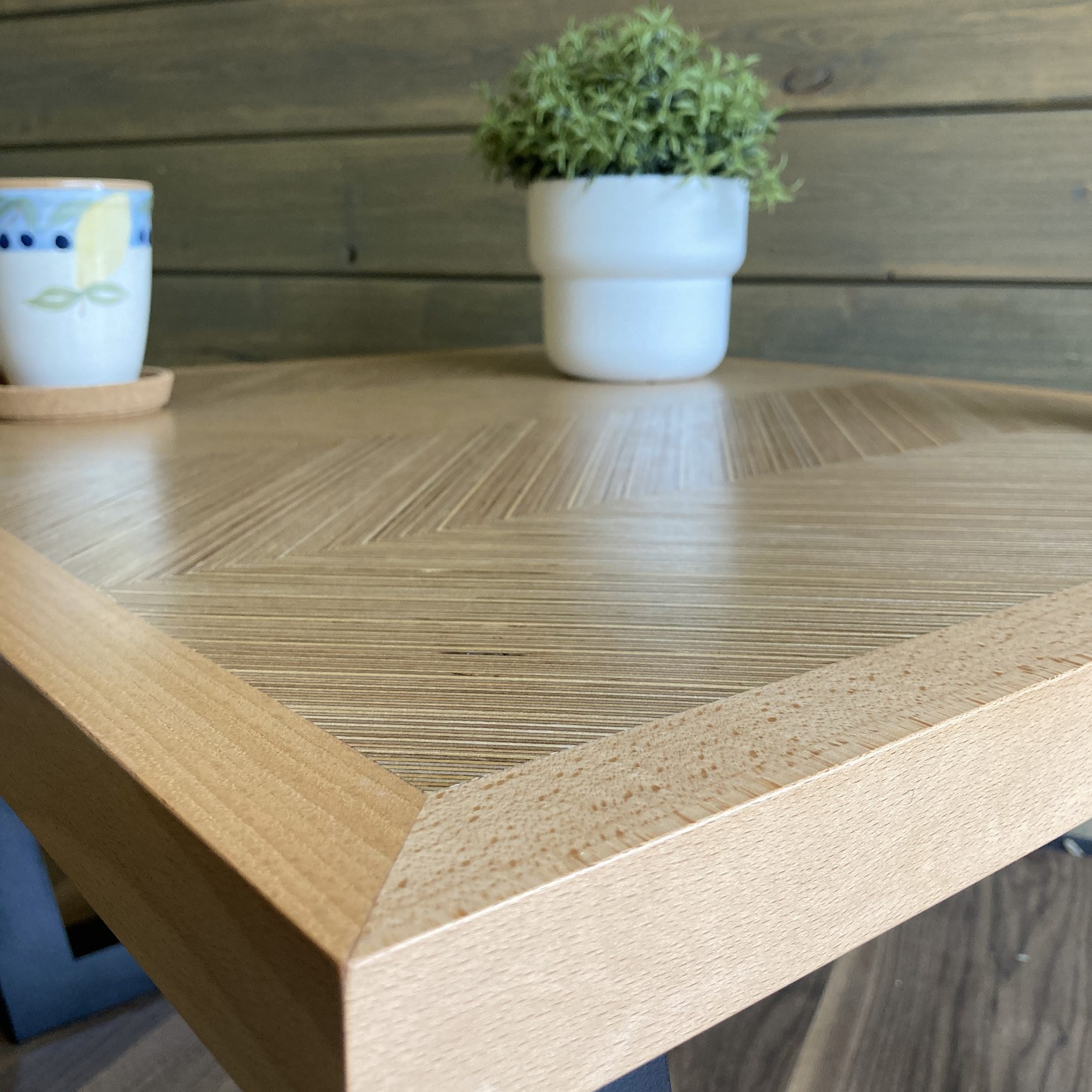 A close corner view of a herringbone patterned birch ply table with solid beech edging. Links to the corresponding product page